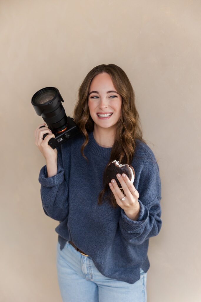 Jenna Richmond smiles and wears a blue sweater and stands in front of a tan wall in a studio with her camera and a whoopie pie in hand.